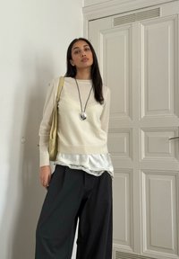 Pulover bej deschis cu mâneci lungi, purtat peste un top gri deschis din satin, cu margine de dantelă. Pantaloni largi negri lejeri și o geantă bej.