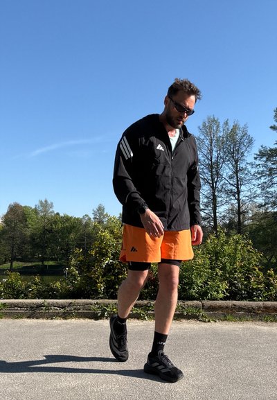 Hombre con chaqueta deportiva negra y pantalones cortos naranjas caminando al aire libre en un día soleado con árboles y arbustos al fondo.