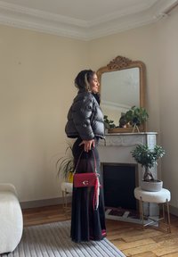Donna in giacca a vento nera e gonna lunga trasparente tiene in mano una borsa rossa, in piedi in una stanza con piante, uno specchio decorato e un camino in marmo.