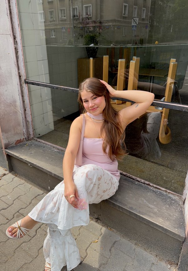 Jeune femme aux longs cheveux bruns assise sur un rebord de trottoir, portant un haut rose, un pantalon blanc en dentelle et des sandales blanches, souriant à la caméra.