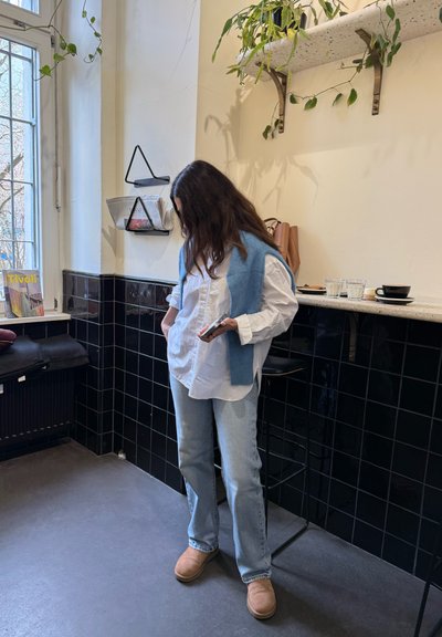 Camisa blanca de botones debajo de un cárdigan azul, jeans azul claro y botas beige. Fondo de azulejos negros con decoración mínima y una ventana.