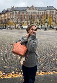 Geantă din piele Jor orange, împletită, cu un mâner superior și o curea de umăr. Geanta prezintă un model distinctiv și un jucărie pufoasă atașată.