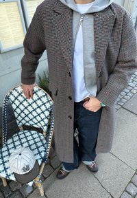 Persona con un abrigo a cuadros y una sudadera con capucha gris, de pie en la acera, sosteniendo una silla tejida azul y blanca con una gorra de béisbol blanca encima.