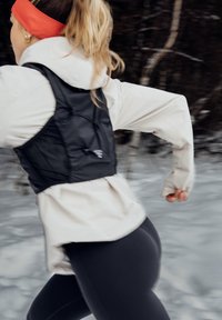 Femme blonde avec un bandeau orange et un gilet noir courant en plein air dans une zone boisée enneigée, portant une veste légère et des leggings noirs.
