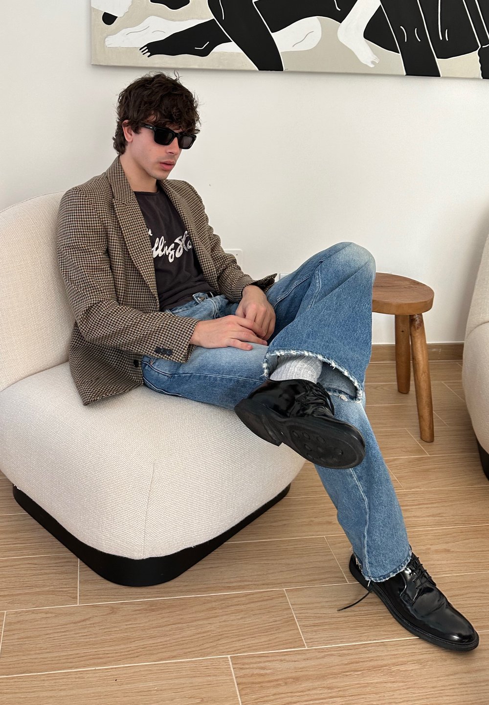 Uomo seduto su un poltrona beige, indossando un blazer a scozzese, t-shirt nera e jeans blu strappati. Scarpe nere e occhiali da sole scuri.