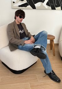 Uomo seduto su un poltrona beige, indossando un blazer a scozzese, t-shirt nera e jeans blu strappati. Scarpe nere e occhiali da sole scuri.
