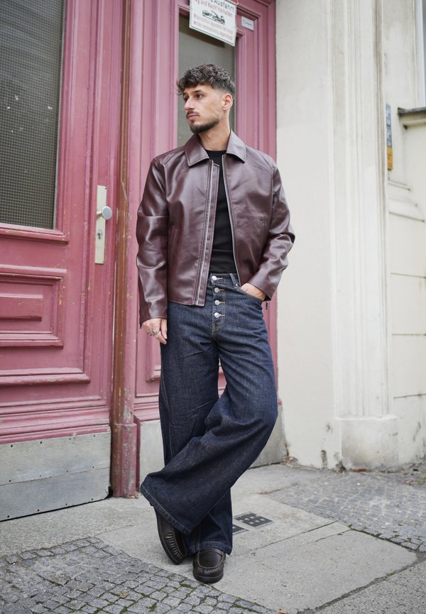 Homme aux cheveux courts et foncés se tient devant une porte rose, portant une veste en cuir marron, une chemise noire, un jean large foncé et des chaussures noires.