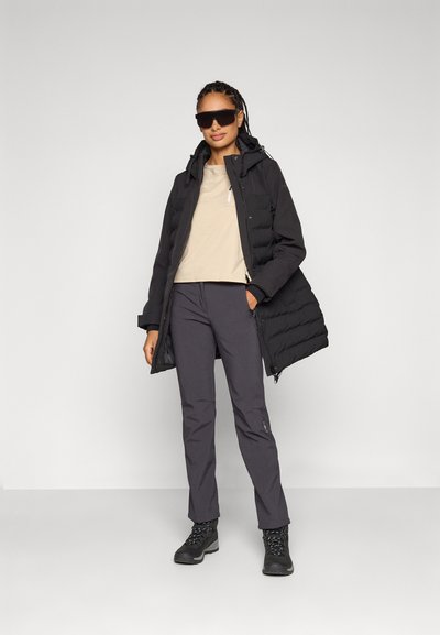 Chaqueta de plumas negra con capucha, top corto beige, pantalones gris oscuro y botas de senderismo negras. La modelo lleva gafas de sol negras de gran tamaño.