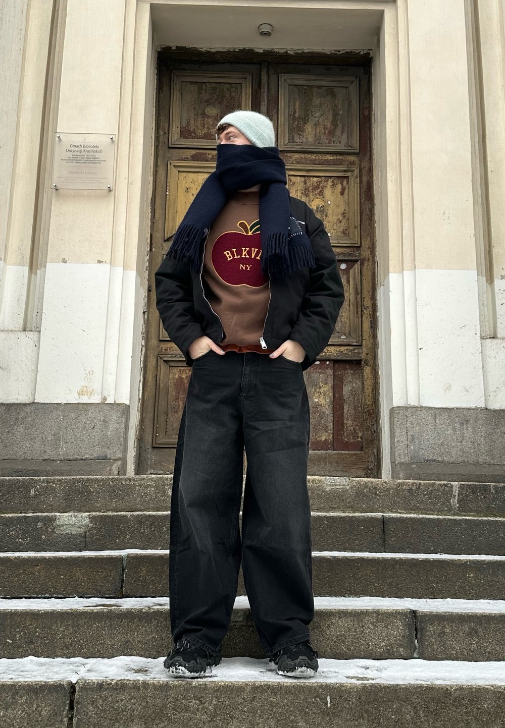 Černá oversized bunda, hnědá mikina s grafikou jablka, tmavé široké kalhoty, námořnický šátek, světle modrý čepice, stojící na kamených schodech.