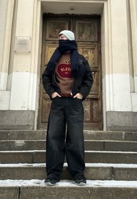 Černá oversized bunda, hnědá mikina s grafikou jablka, tmavé široké kalhoty, námořnický šátek, světle modrý čepice, stojící na kamených schodech.