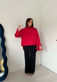 Röd oversized långärmad topp med hög krage, i kombination med svarta plisserade byxor och svarta sandaler. Slät tygtextur.