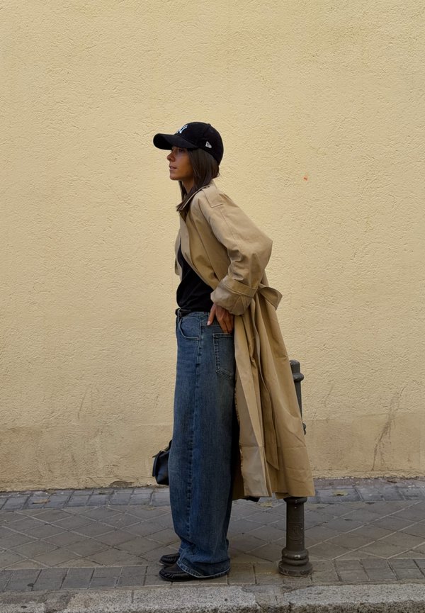 Une femme portant une casquette noire, un trench beige, un haut noir et un jean à jambes larges se tient près d’un poteau, contre un mur beige.