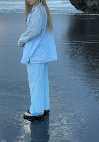 Jasnoniebieska, oversize koszula na guzik i dopasowane szerokie spodnie wykonane z lekkiego materiału, w połączeniu z czarnymi klapkami, stojąc na mokrym piasku.