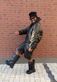 Fekete bőr bombázó dzseki fekete felső fölött, fekete bőr rövidnadrággal és térdig érő fekete gumicsizmával, texturált talppal.