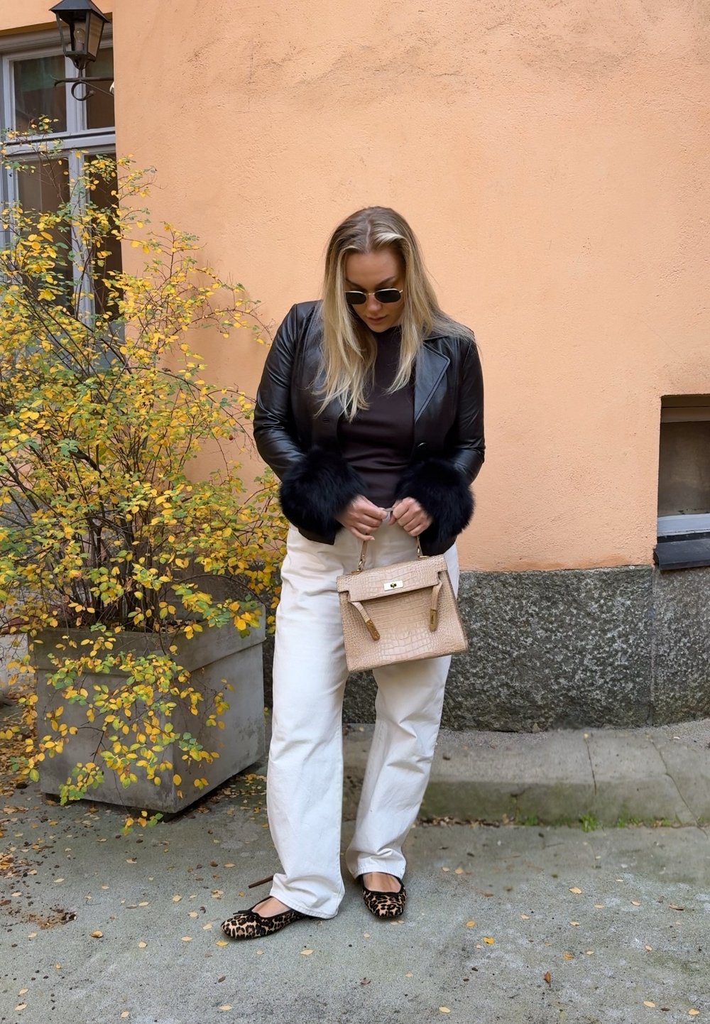 Giacca in pelle nera con polsini in pelliccia, top marrone, pantaloni bianchi a gamba larga, scarpe basse con stampa leopardata e una piccola borsa beige testurizzata.