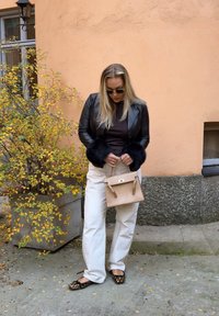Giacca in pelle nera con polsini in pelliccia, top marrone, pantaloni bianchi a gamba larga, scarpe basse con stampa leopardata e una piccola borsa beige testurizzata.