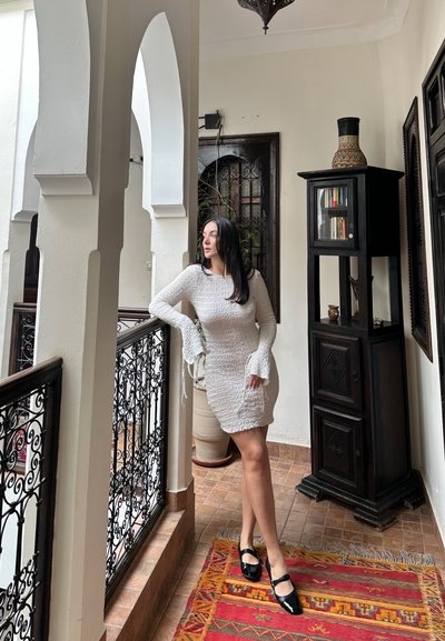 Vestido blanco de manga larga y textura con puños acampanados. Zapatos negros de charol estilo Mary Jane. Ambiente interior con una alfombra estampada y muebles de madera.