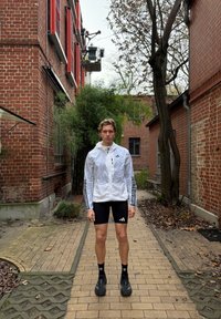 Veste blanche zippée avec des bandes noires, short noir avec un logo Adidas, chaussettes noires et chaussures foncées. Chemin extérieur pavé en arrière-plan.