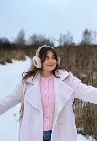 Jeune femme portant un manteau blanc, un pull rose et des cache-oreilles, debout les bras écartés sur un chemin enneigé avec des buissons secs en arrière-plan.