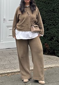 Kobieta ubrana w beżowe szerokie spodnie, dopasowany hoodie na białej koszuli, beżowe buty i beżową torebkę, stojąca na wybrukowanym chodniku obok żywopłotu.