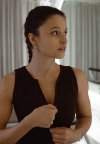 Žena s vrkočmi vo vrkočoch v tmavom tielku bez rukávov zapína zips, pozerá sa bokom v mäkko osvetlenej vnútornej miestnosti s závesmi.