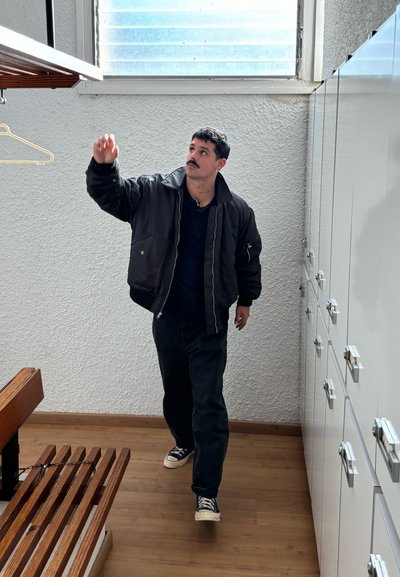 Persona vestida con una chaqueta bomber negra y pantalones oscuros, alcanzando una estantería de madera en una habitación con paredes blancas y suelo de madera clara.