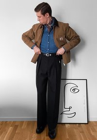 Άνδρας με καφέ σακάκι, μπλε πουκάμισο και μαύρο παντελόνι ψηλής μέσης, που ρυθμίζει τη ζώνη του, με μινιμαλιστική γραμμή προσώπου, ακουμπισμένος σε λευκό τοίχο.