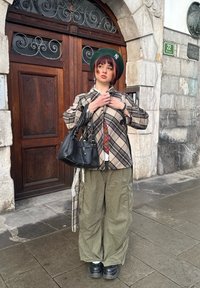 Νεαρό άτομο που φορά πράσινο μπέρε, καρό σακάκι, παντελόνι cargo και κρατάει μαύρη τσάντα, στέκεται στο δρόμο μπροστά από ξύλινη πόρτα.