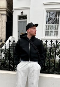 En sort hættetrøje med lynlås parret med lysegrå sweatpants. Hættetrøjen har en h�ætte og et flettet bånd, sat mod en hvid bygning i baggrunden.