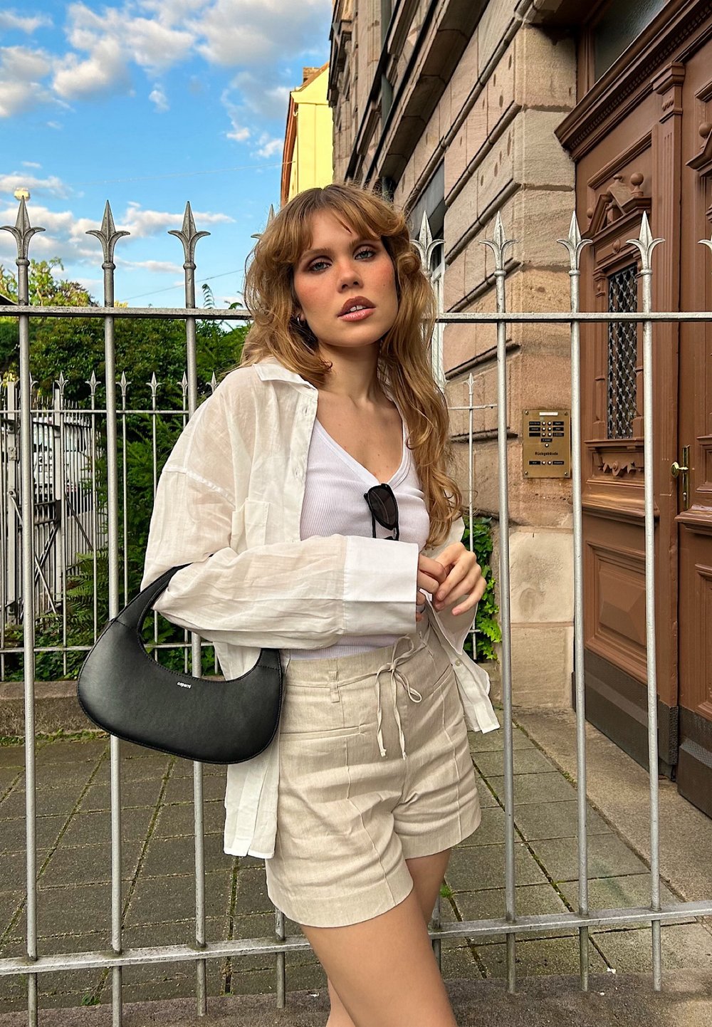 Camicia oversized in lino bianco, canotta bianca, pantaloni beige e borsa a tracolla nera. Sullo sfondo si vede una recinzione in ferro battuto e un edificio in pietra.