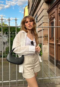 Camicia oversized in lino bianco, canotta bianca, pantaloni beige e borsa a tracolla nera. Sullo sfondo si vede una recinzione in ferro battuto e un edificio in pietra.