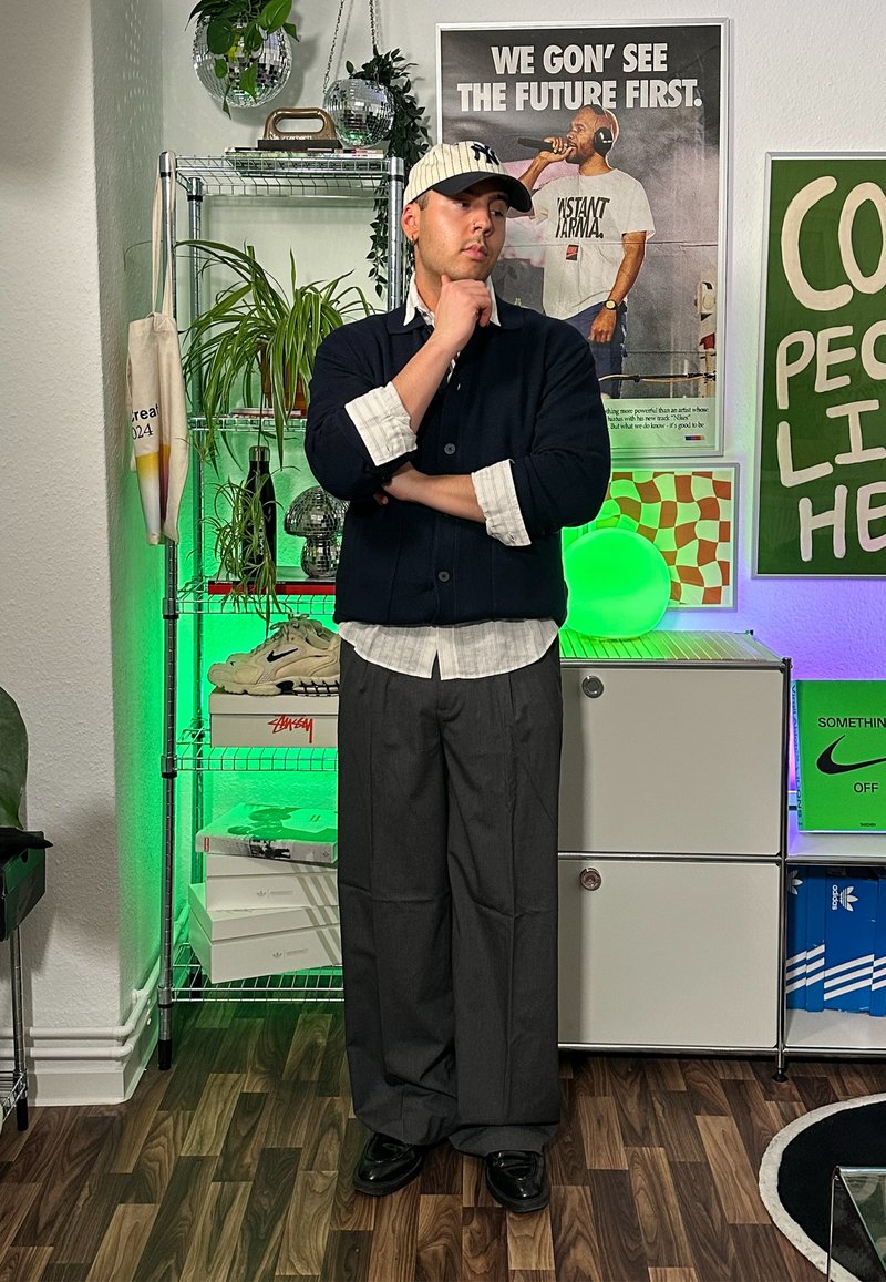 Navy knitted cardigan, white button-down shirt, grey wide-leg trousers, black shoes. The background includes green lighting and various decorative items.