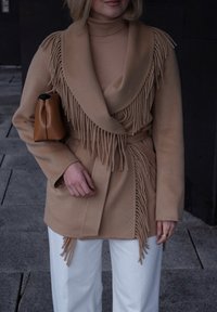 Manteau enveloppant en laine beige avec une bordure à franges dégradées, associé à un col roulé ajusté beige clair et un pantalon blanc à jambes larges, tenant un petit sac marron.