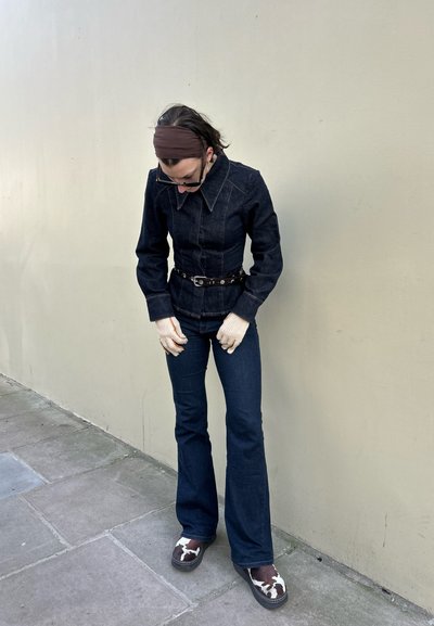 Chaqueta de mezclilla oscura con cuello punta, cintura ceñida y cinturón negro. Vaqueros oscuros ajustados y calzado con estampado complementan el conjunto.