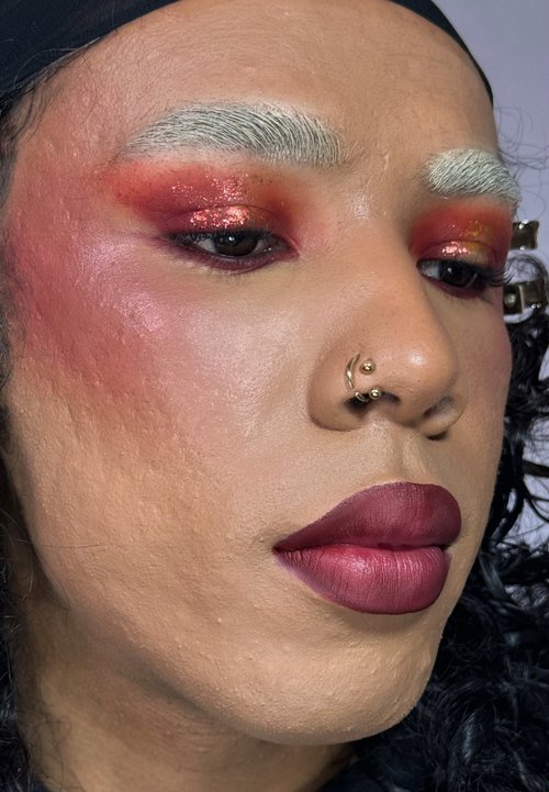 Närbild på ett ansikte med texturerad hud, djärva burgunderröda läppar och dramatisk ögonmakeup med inslag av rött och guldglitter. Framträdande silverfärgade ögonbryn.