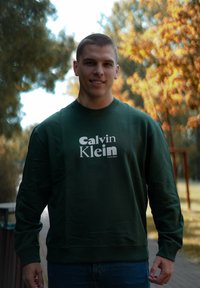 Grønn hettegenser med ribbede mansjetter og halslinning. Har "Calvin Klein" logo i hvit skrift på brystet. Brukt utendørs, omgitt av trær.