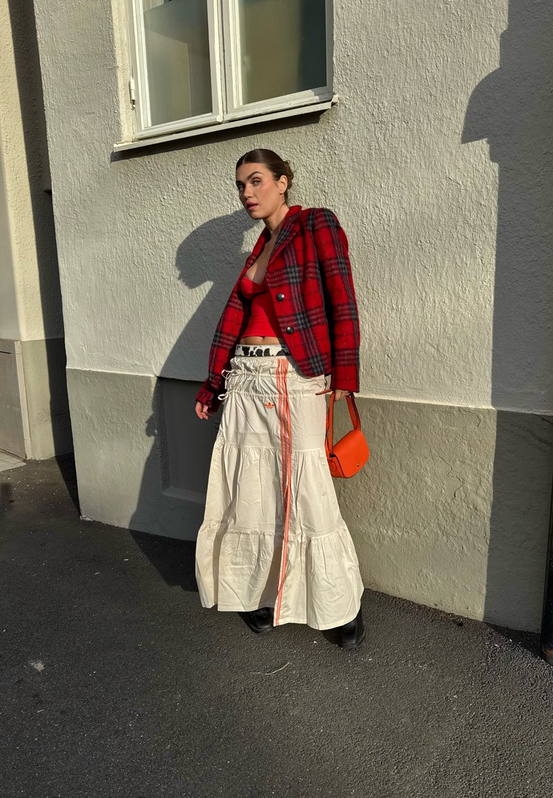 Blazer xadrez vermelho, top justo vermelho, saia creme em camadas com cordão laranja, e pequena mala laranja. Botas pretas. Sombra na parede.