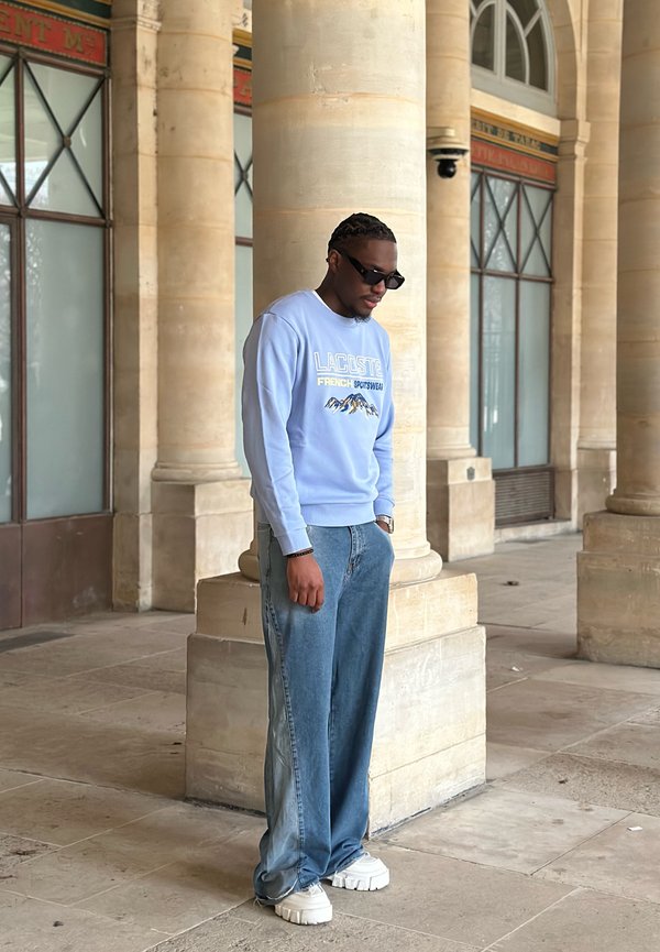 Άνδρας που φοράει ανοιχτό μπλε φούτερ Lacoste, φαρδιά τζιν, λευκά αθλητικά παπούτσια και μαύρα γυαλιά ηλίου, στέκεται κοντά σε πέτρινες κολόνες σε καλυμμένο πεζόδρομο.