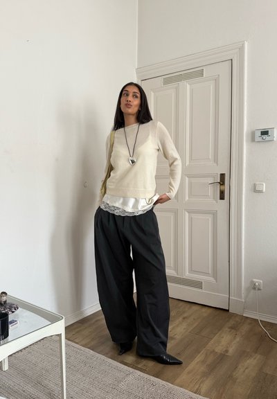 Suéter crema superpuesto sobre una blusa blanca de satén con ribete de encaje, combinado con pantalones negros holgados y zapatos negros puntiagudos. Cabello largo y oscuro.