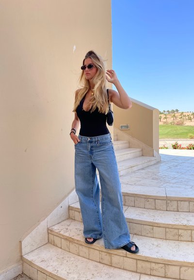 Bodysuit negro sin mangas, jeans de pierna ancha color azul claro, gafas de sol negras y sandalias negras, de pie en una escalera de mármol con paredes beige.