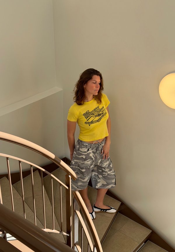 Mujer con cabello rizado, camiseta gráfica amarilla y pantalones cortos de camuflaje, parada tranquilamente en una escalera de caracol con baldosas al lado de una luz de pared redonda.