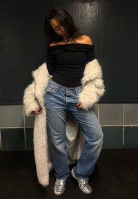 Svart ribbete off-shoulder topp, hvit fluffy lang jakke, løse blå denimjeans, sølv sneakers med teksturert design, og et lagdelt halskjede.