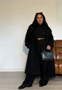 Mustat trench-takit mustan poolopaidan ja leveälahkeisten housujen päällä, tanvärinen vyö ja pieni musta käsilaukku, jossa on kultaiset yksityiskohdat.