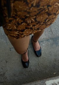 Schwarze Lackleder-Ballettschuhe mit quadratischen Spitzen und einem dünnen Knöchelriemen. Der Hintergrund zeigt eine strukturierte Oberfläche und einen Rock mit Blumenmuster.