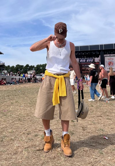 Pantalones cortos color tostado con un suéter amarillo anudado, camiseta blanca, gorra marrón y botas amarillas. Collares en capas adornan el cuello.