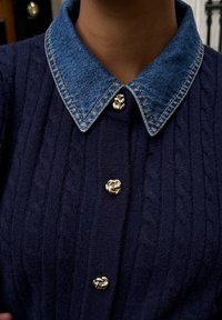 Granatowy sweter z dzianiny z kołnierzem z denimu i ozdobnymi złotymi guzikami. Materiał ma wzór warkoczowego splotu.