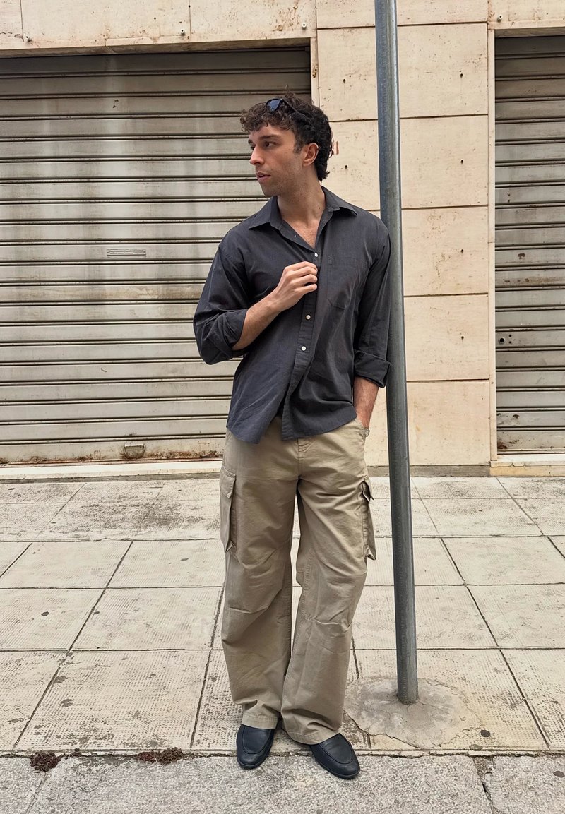Homme portant une chemise foncée et un pantalon cargo beige se tient sur le trottoir à côté d'un poteau métallique et de volets métalliques fermés, regardant sur le côté.