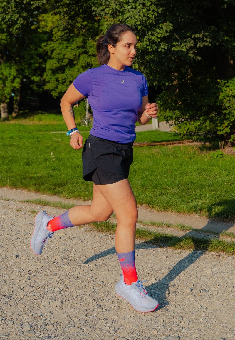 Uma mulher a correr numa trilha de gravilha, vestindo uma camisola roxa, calções pretos e meias roxo-avermelhadas, com árvores verdes ao fundo.