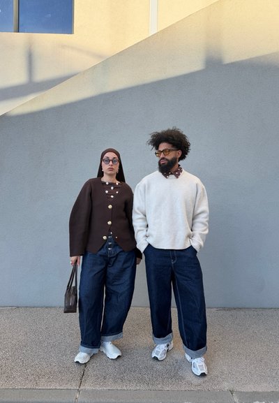 Dos personas de pie una al lado de la otra, vistiendo jeans sueltos y suéteres casuales, ambas con gafas de sol y zapatillas blancas, frente a una pared gris.