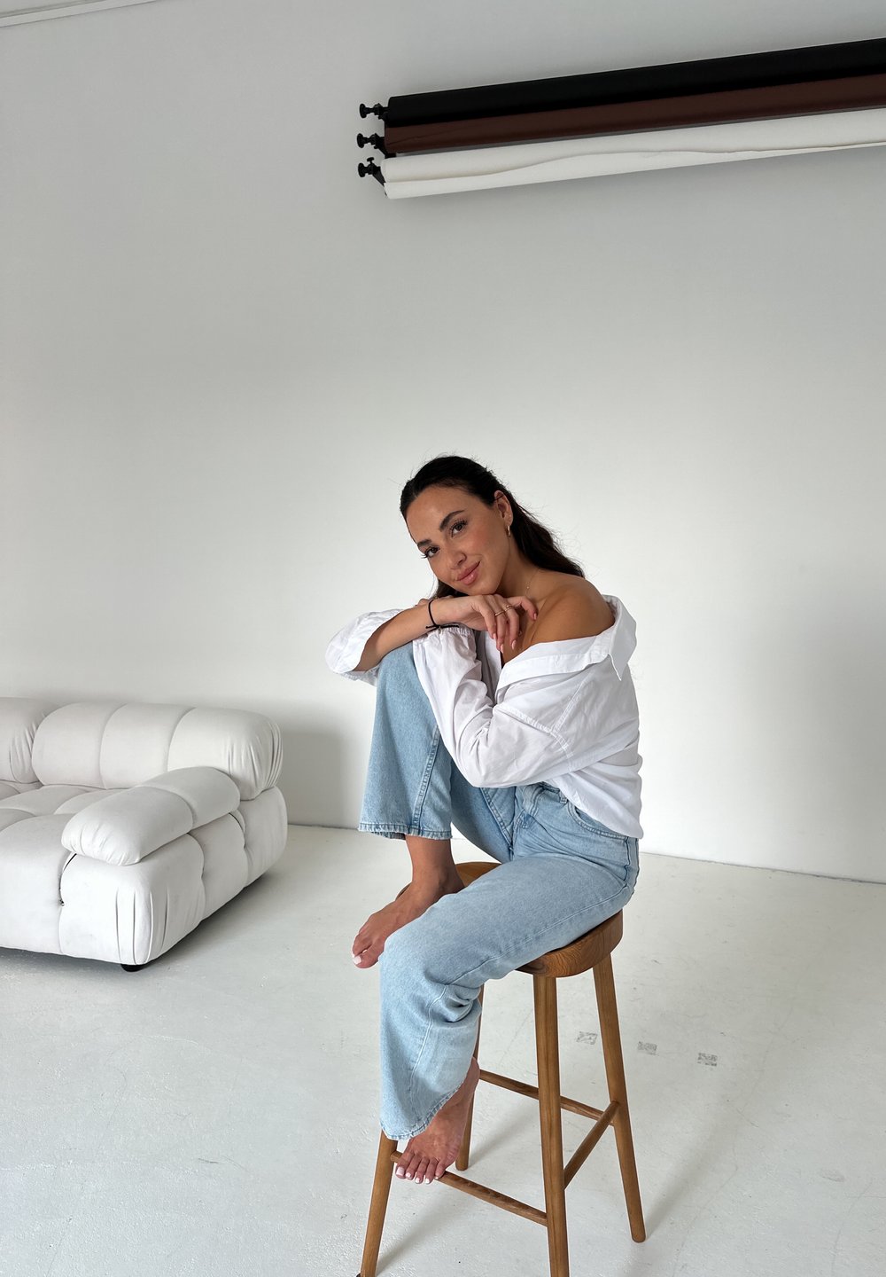 Witte oversized shirt, lichtblauwe jeans, zittend op een houten kruk. Pluche witte bank op de achtergrond, minimalistische setting met neutrale tinten.